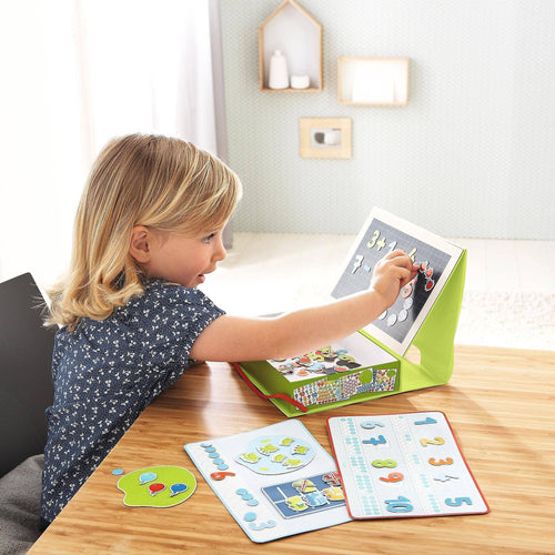 A young child playing with the 1, 2 Numbers & You Magnetic 158 Piece Game Box, using magnetic numbers and colorful educational boards on a wooden table in a bright, cozy room.