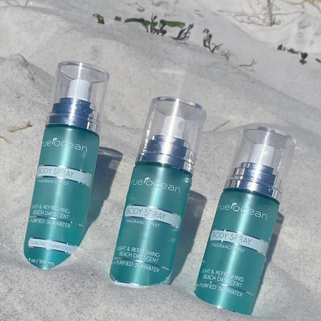 Three bottles of True Ocean - True Ocean: Body Spray - 3.4oz. with teal packaging and clear caps arranged on white sand, evoking a fresh, beachy vibe.