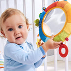 Baby holding a colorful Rainbow Discovery Mirror with soft fabric textures and attached sensory toys, designed for engaging and educational play in a cozy nursery setting.