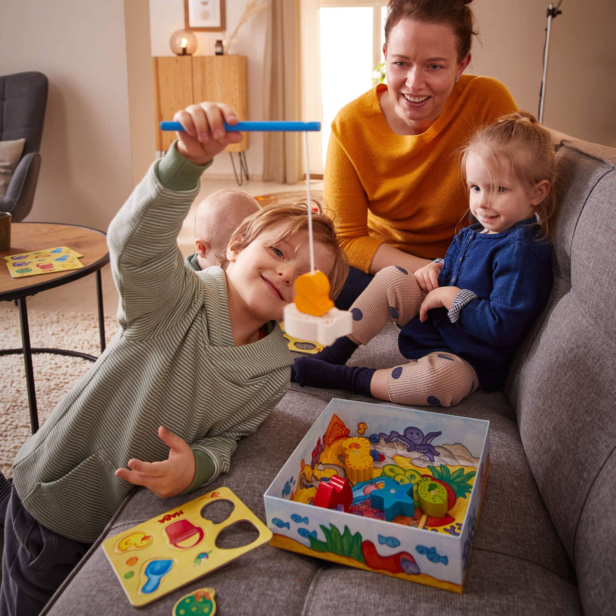 A young boy playing "My Very First Games - Here, Fishy, Fishy! Magnetic Game" with a fishing rod toy while a smiling woman and a toddler watch on a cozy couch, promoting stylish kids clothing and educational toys in a curated children’s boutique.