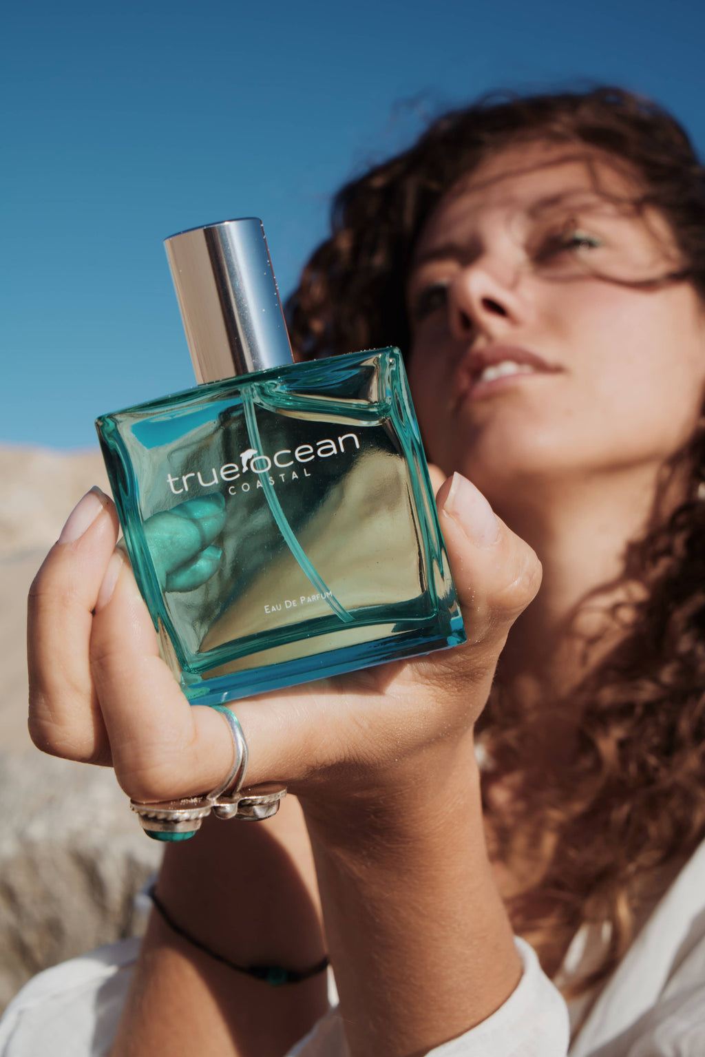 Woman holding a bottle of True Ocean - Coastal - a beach perfume outdoors, with a clear blue sky and sunlit natural background, embodying calm and fresh coastal vibes.