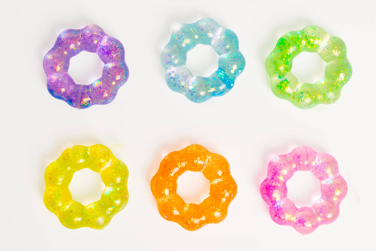 Six flower-shaped translucent resin rings with scalloped edges and embedded glitter, arranged in two rows of three on a plain white background. The rings come in purple, blue, green, yellow, orange, and pink.
