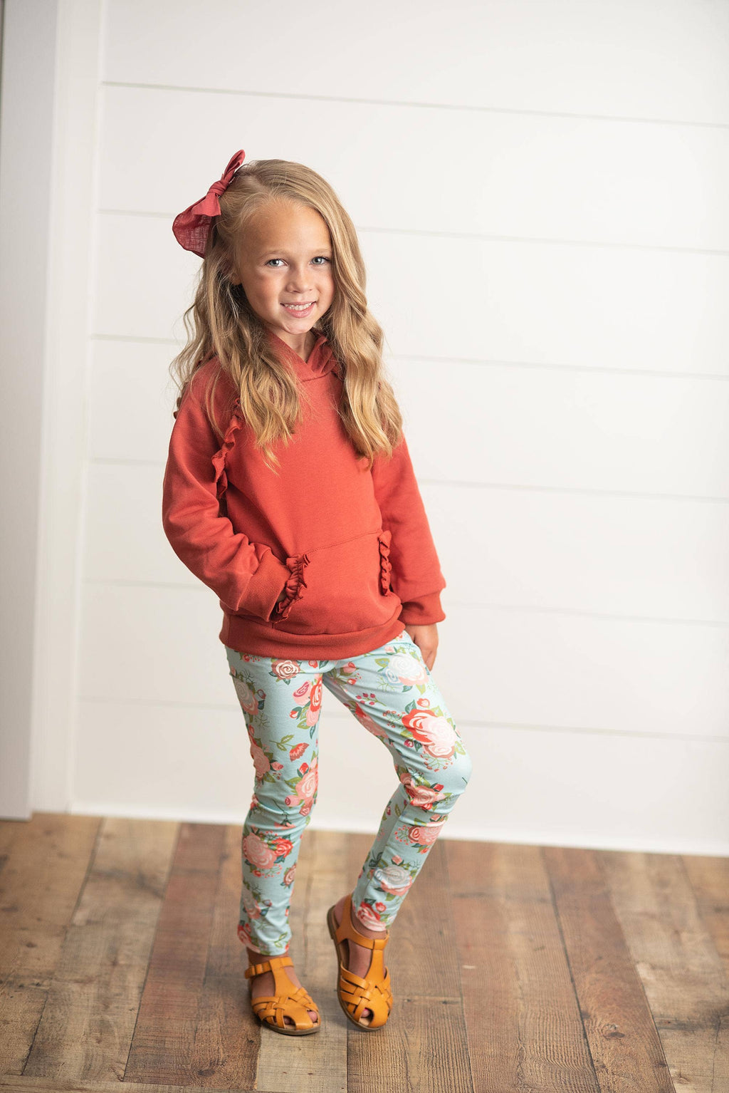 A young girl with blonde hair wearing a coral orange ruffle-trimmed hoodie, light blue leggings with a pink and white pattern, and orange shoes, standing against a white shiplap wall and wood floor.