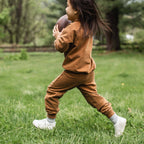 Child in brown football outfit holding a football on grass.