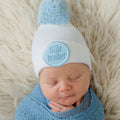 Newborn boy wearing the ilybean - White Little Brother Newborn Boy Hospital Hat Handmade Pom in soft white knit with a blue pom, wrapped in a cozy blue blanket against a fluffy backdrop, stylish essentials for little ones.