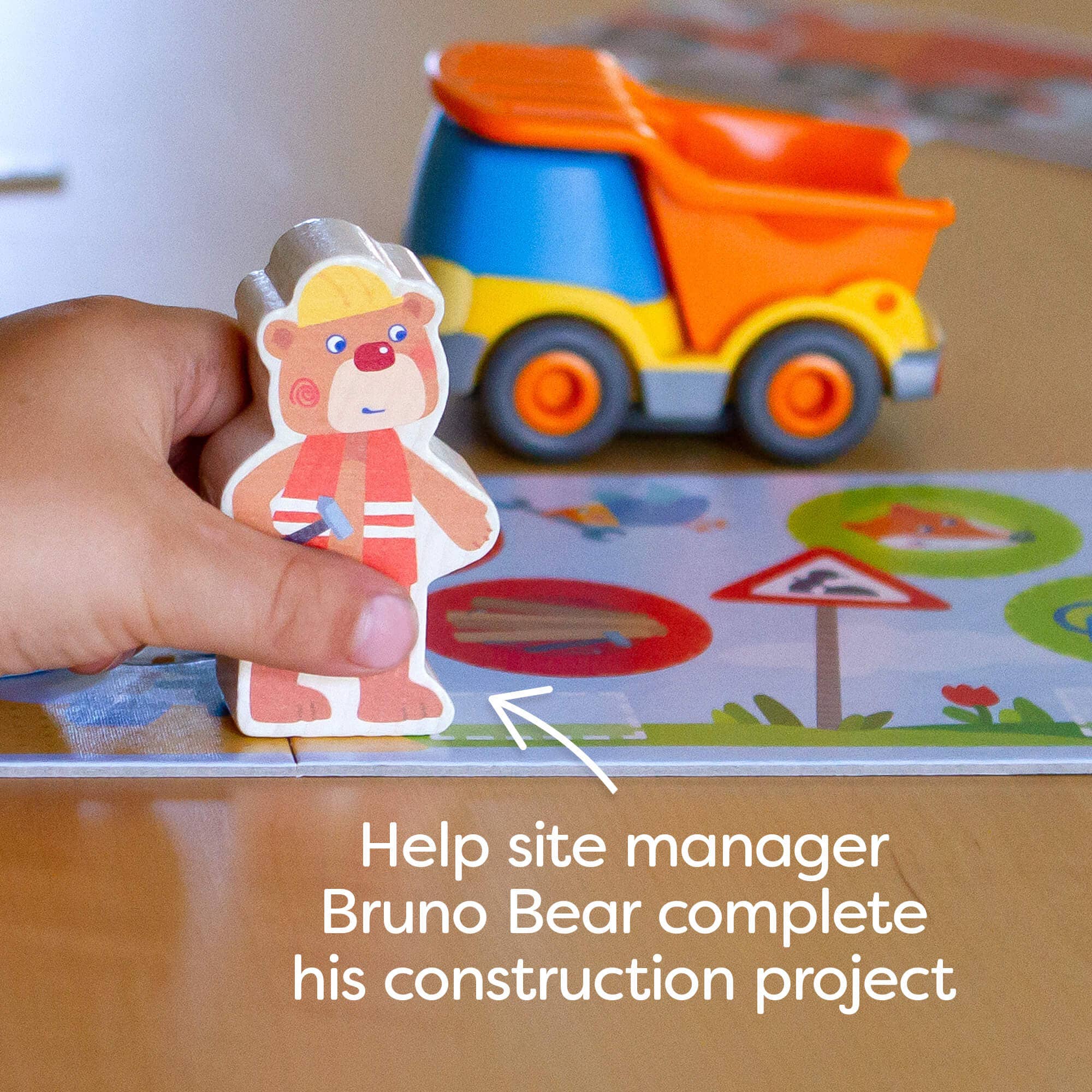 Child's hand holding a Bruno Bear figure wearing a construction hat and vest from My Very First Games - Building Site, with a toy dump truck and construction signs in the background.