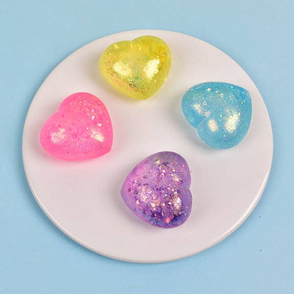Four heart-shaped glittery squishies in pink, yellow, blue, and purple, arranged on a small white circular plate against a light blue background.