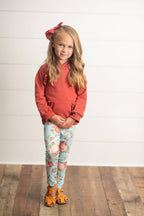A young girl stands against a white paneled wall on a wooden floor, wearing a rust-orange ruffle-sleeve hoodie, light blue floral leggings, and orange sandals with a matching hair bow.