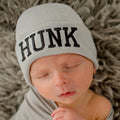 Sleeping newborn wearing a soft gray hat with the word "HUNK" embroidered, showcasing the ilybean - HUNK - Newborn and Baby Hospital Hat: Gray Hat from a curated children’s boutique.