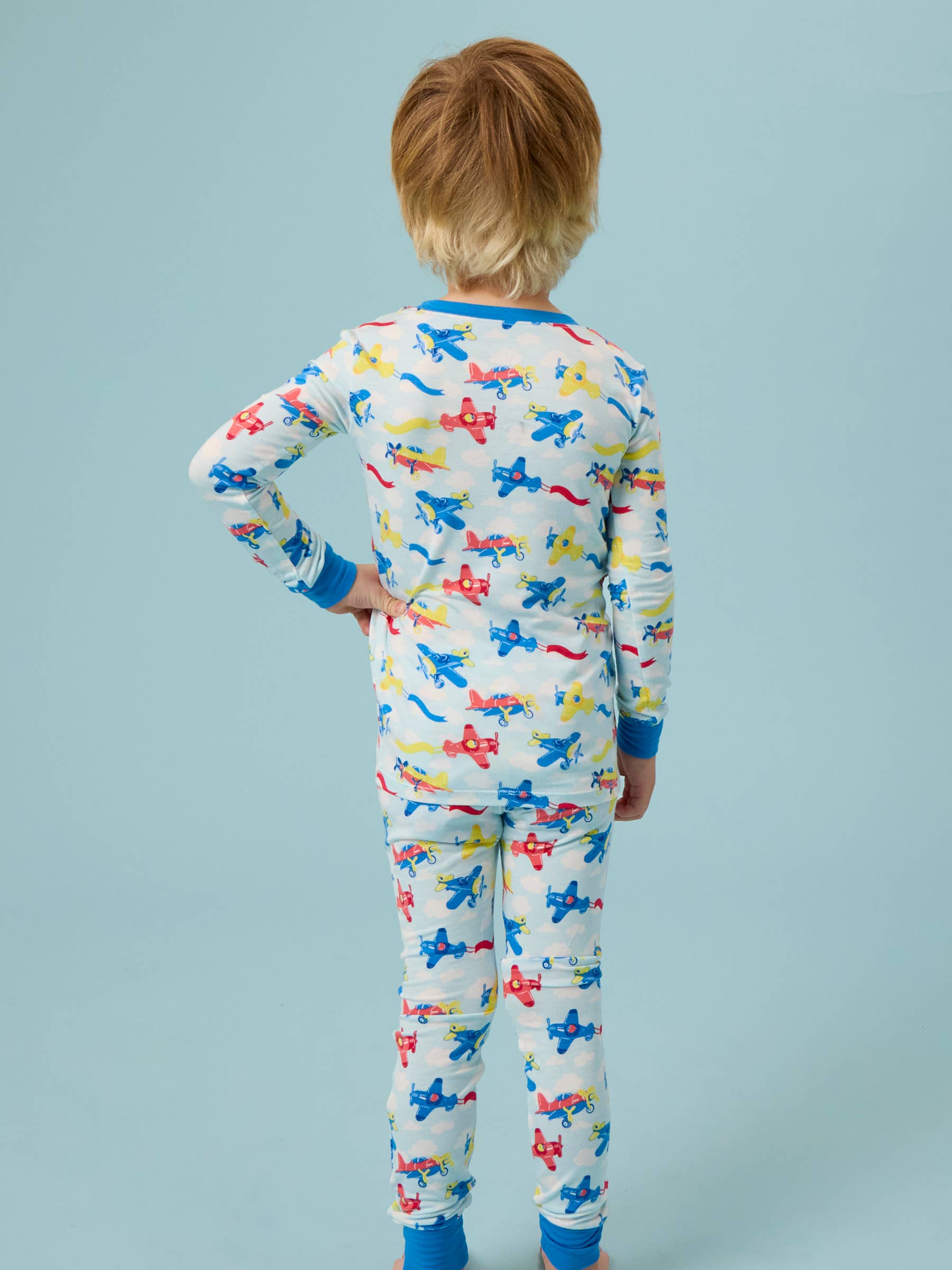Child wearing RuffleButts + RuggedButts - SoftSnooze™ Kids Bamboo Viscose Plane Parade Long Sleeve Pajama Set featuring colorful airplanes on a soft neutral background, cozy and breathable for bedtime comfort.