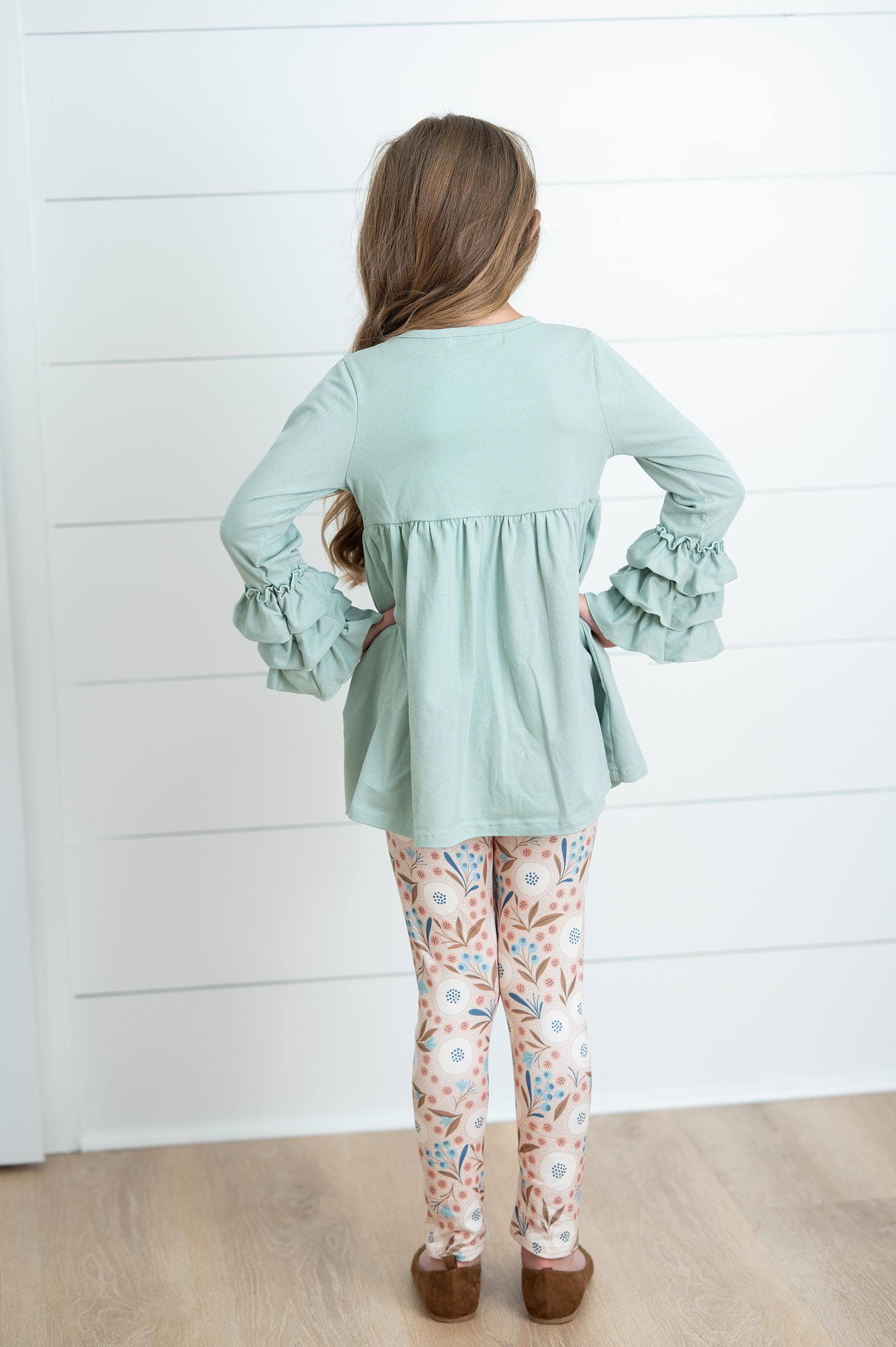 Person wearing a light green top with ruffled sleeves and floral leggings against a white wall.