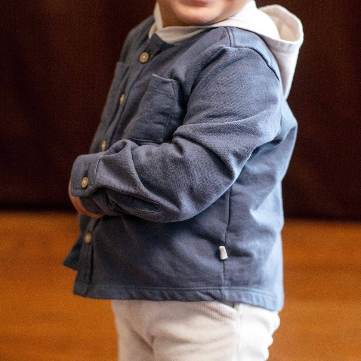 A toddler wearing a blue sweatshirt jacket with a white hood and white pants, standing in an indoor setting with a wooden floor and dark brown background.
