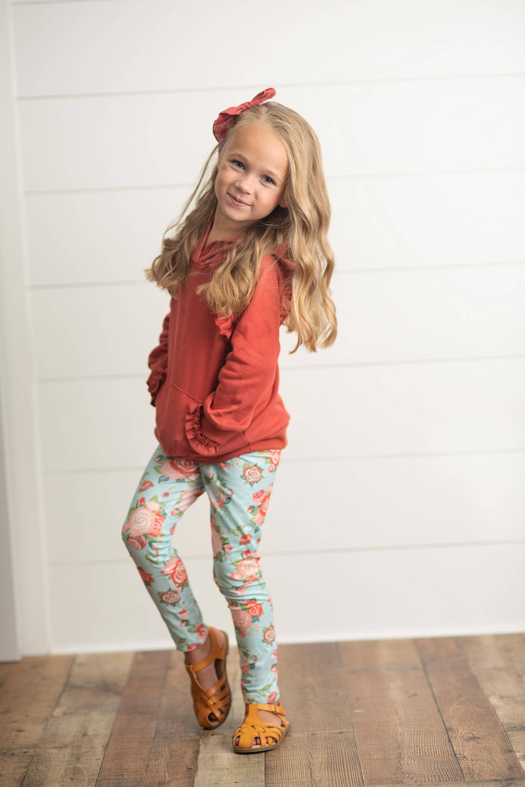A young girl with blonde hair wearing a rust-orange ruffle-hem hoodie, light blue floral leggings, and brown sandals, standing against a white shiplap wall.