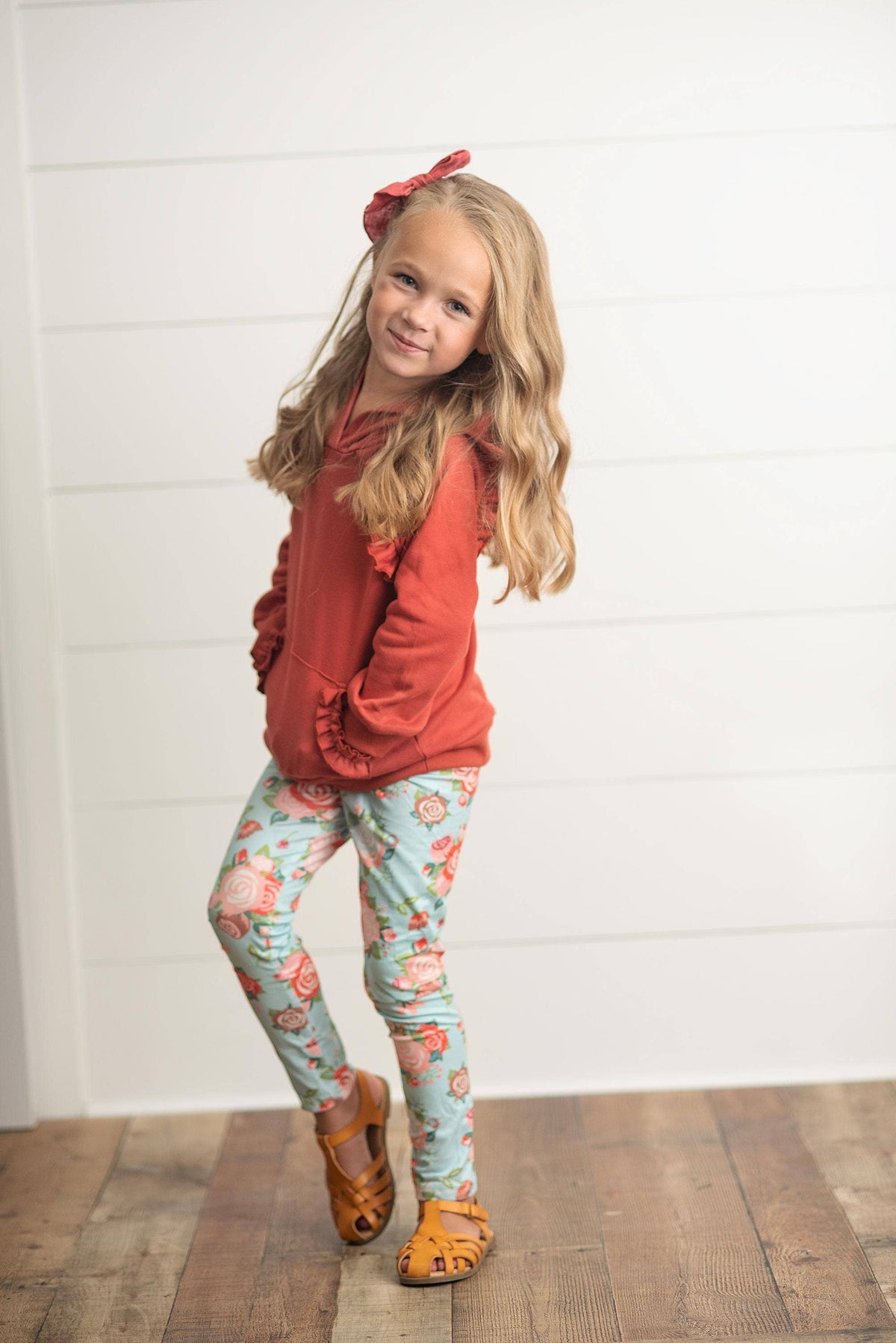 A young girl with blonde hair wearing a rust-orange ruffle-hem hoodie, light blue floral leggings, and brown sandals, standing against a white shiplap wall.