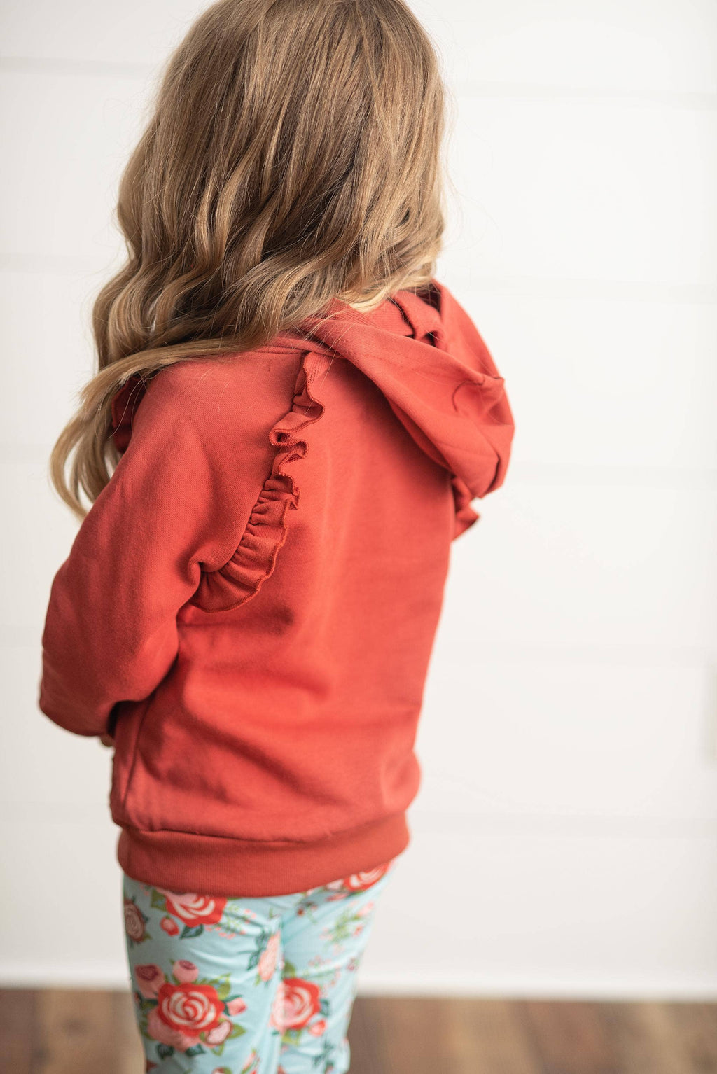 A child viewed from behind wearing a rust-orange hooded sweatshirt with ruffle detailing on the hood and sleeve, paired with light blue floral leggings, against a white wall background.