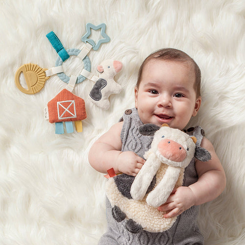 Baby lying on a soft cream-colored blanket holding a plush cow toy, smiling next to a fabric toy set with a barn, star-shaped teether, and cow, featuring the Itzy Ritzy - Bitzy Busy Gift Set™.