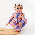 Smiling baby wearing a Kiki + Lulu - Pink Dino Convertible Footie with colorful dinosaur and nature prints, sitting on a white floor and wearing a white bow headband.