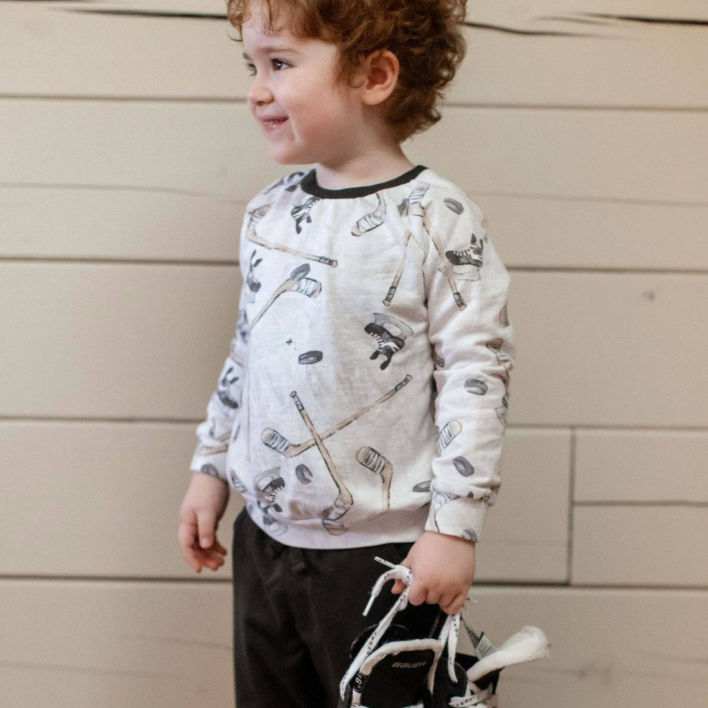Toddler wearing Burt's Bees Baby - Hockey Season Sweatshirt & Pants Set with hockey-themed print, holding a pair of ice skates, styled in soft neutral colors perfect for modern baby boutique collections.