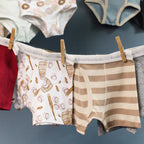 A close-up of several pairs of children's underwear hanging on a dark blue wall with wooden clothespins. The center pair is white with a beige and brown pastry print, next to a beige and cream striped pair.