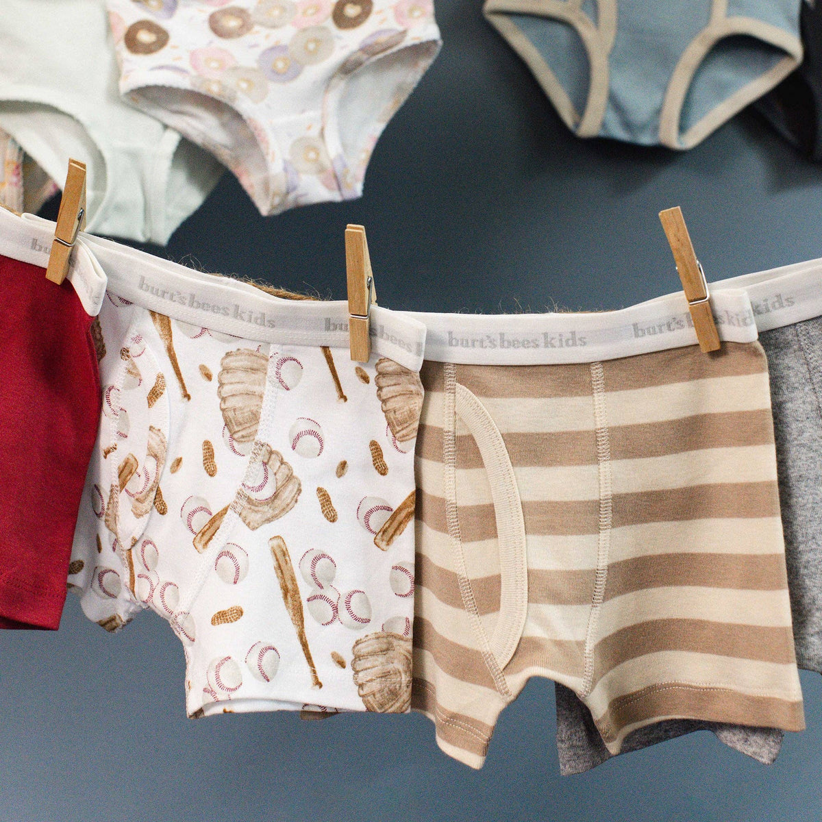 A close-up of several pairs of children's underwear hanging on a dark blue wall with wooden clothespins. The center pair is white with a beige and brown pastry print, next to a beige and cream striped pair.