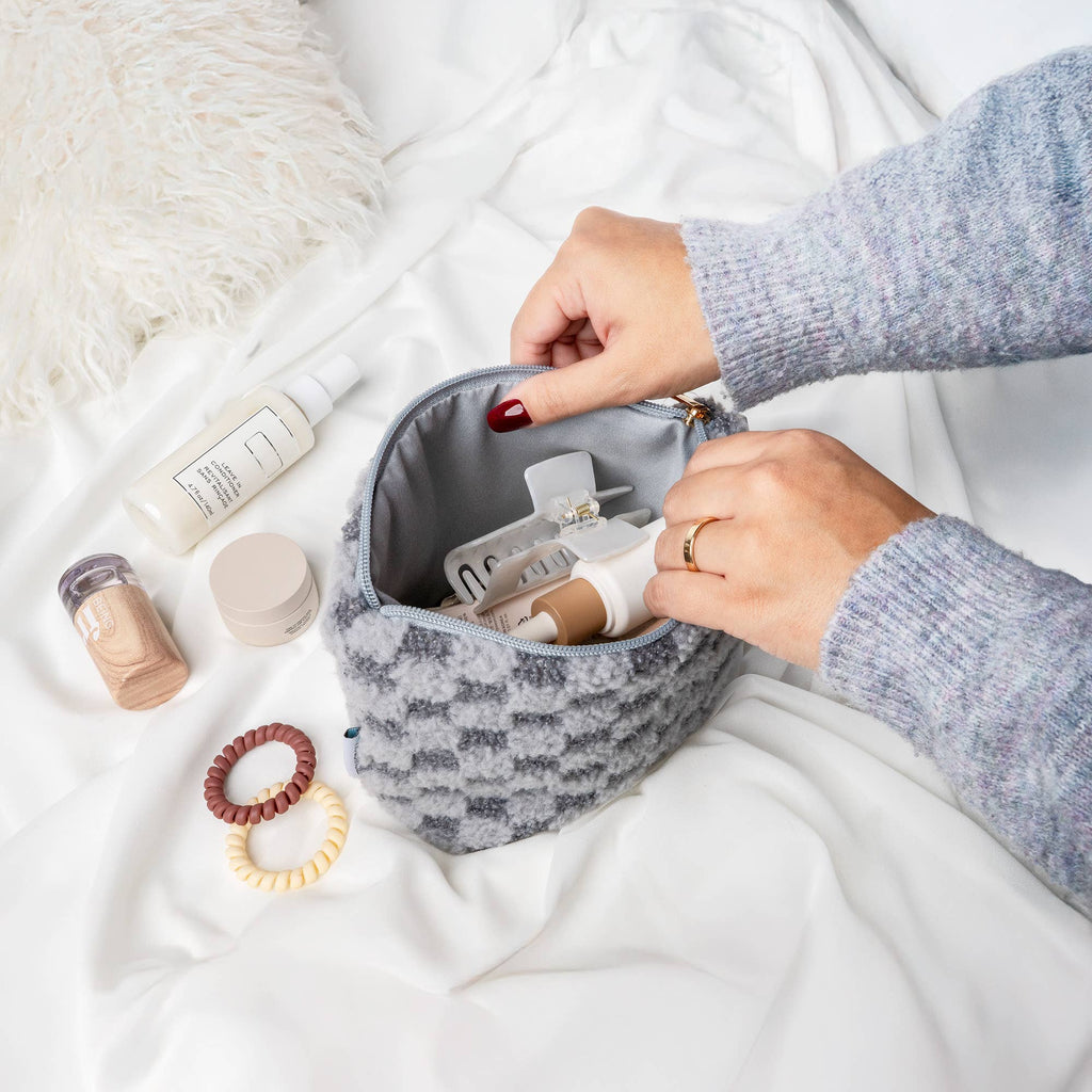 Hands packing neutral baby and mom essentials into a soft textured gray pouch, showcasing the Hello Mello - Good Stuff Essentials Bag in a cozy, modern nursery setting.