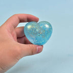 A hand holding a small, heart-shaped, translucent blue object filled with shimmering gold glitter against a solid light blue background.