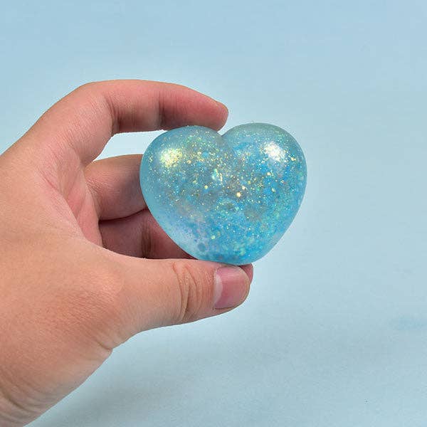 A hand holding a small, heart-shaped, translucent blue object filled with shimmering gold glitter against a solid light blue background.