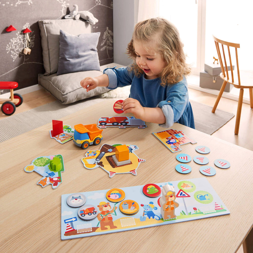A young girl playing with the colorful and interactive pieces of "My Very First Games - Building Site" on a wooden table in a cozy, softly lit playroom designed for imaginative learning and sensory development.