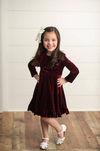 Young girl in a burgundy velvet dress with a white bow standing on a wooden floor.