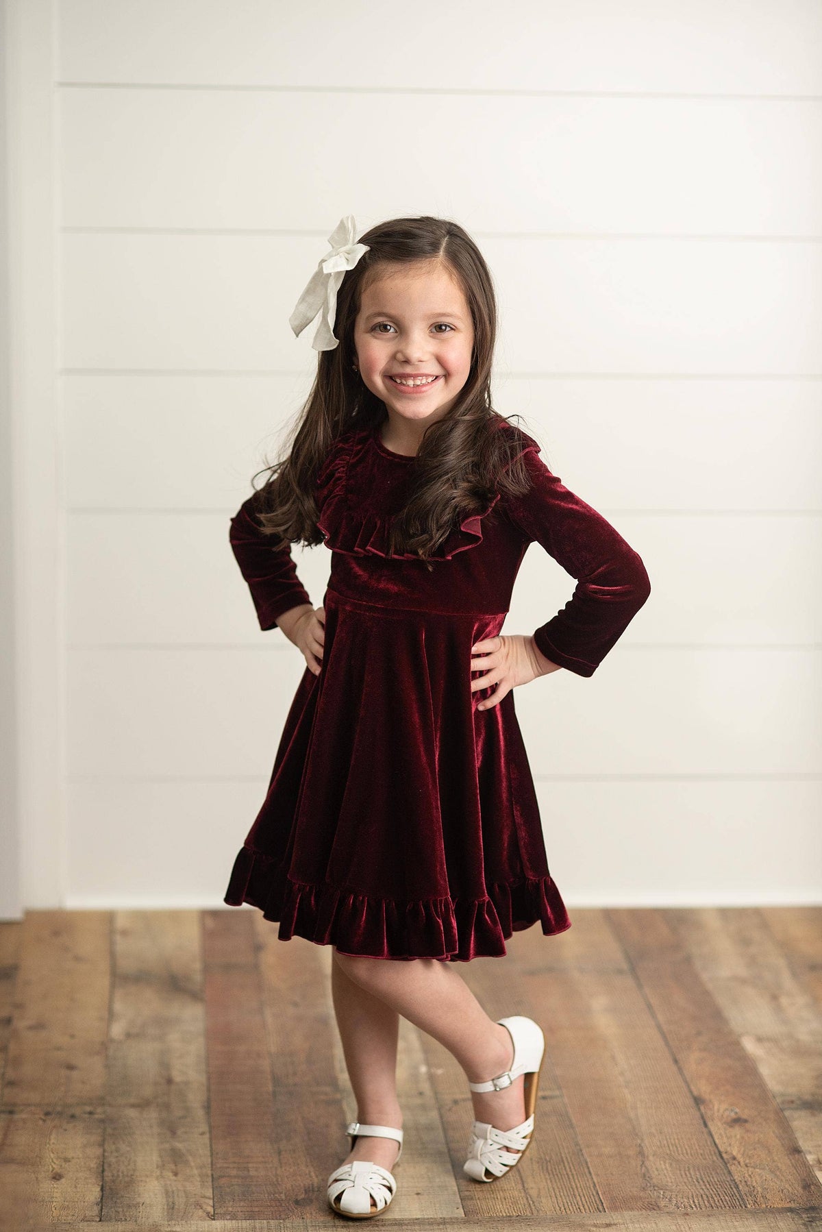 Young girl in a burgundy velvet dress with a white bow standing on a wooden floor.