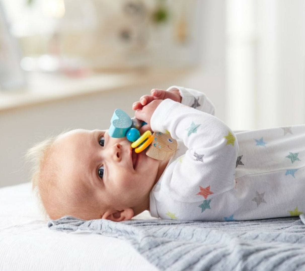 Baby lying on a soft gray blanket holding and chewing on the Rattlefish Wooden Baby Rattle, featuring colorful beads and natural wood, perfect for sensory play and soothing teething.