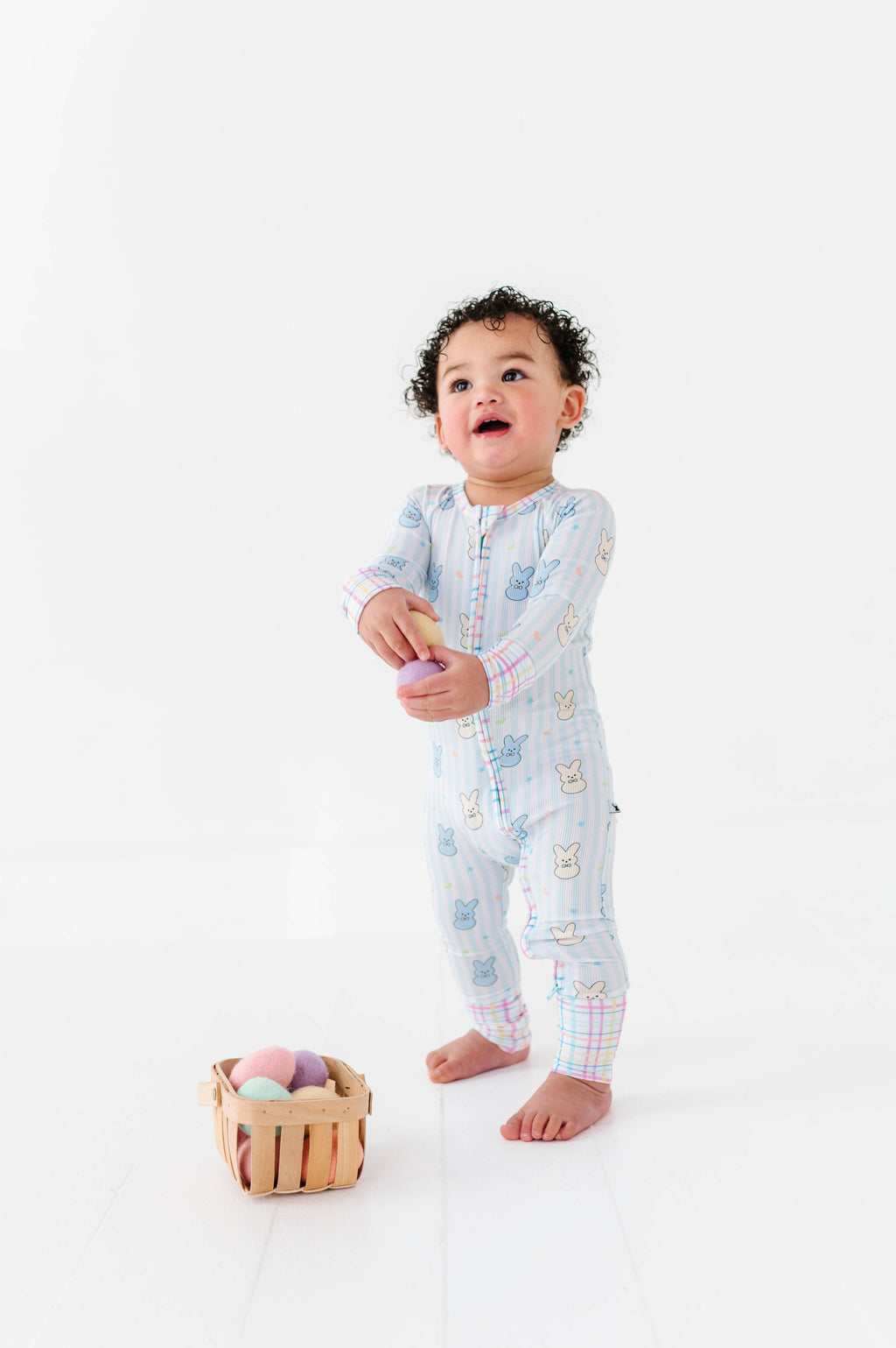 Toddler wearing Kiki + Lulu - Peeps + Sleeps Convertible Footies with pastel bunny prints and plaid cuffs, standing barefoot near a small wooden basket of colorful soft eggs.