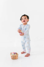 Toddler wearing Kiki + Lulu - Peeps + Sleeps Convertible Footies with pastel bunny prints and plaid cuffs, standing barefoot near a small wooden basket of colorful soft eggs.