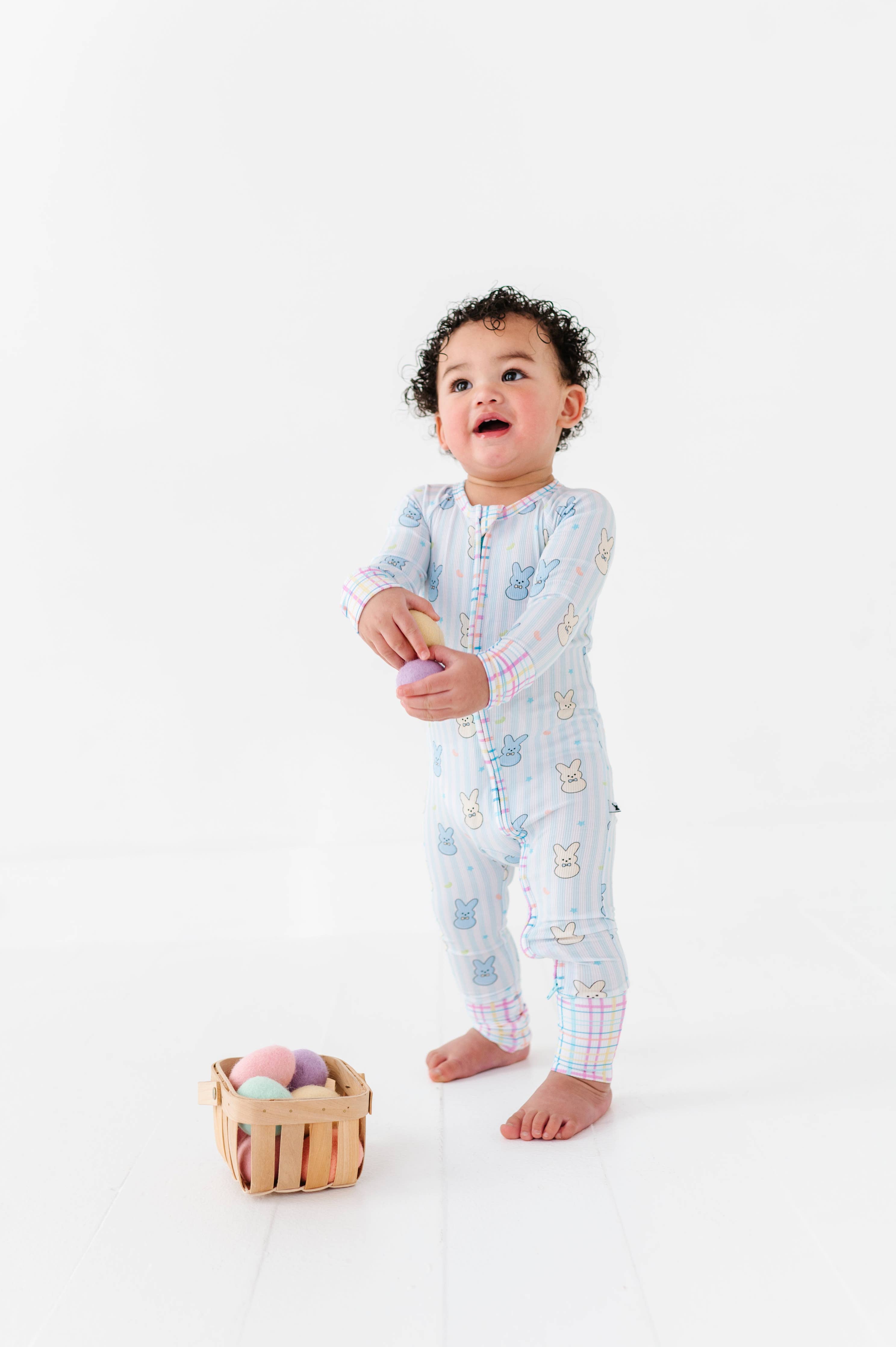 Toddler wearing Kiki + Lulu - Peeps + Sleeps Convertible Footies with pastel bunny prints and plaid cuffs, standing barefoot near a small wooden basket of colorful soft eggs.