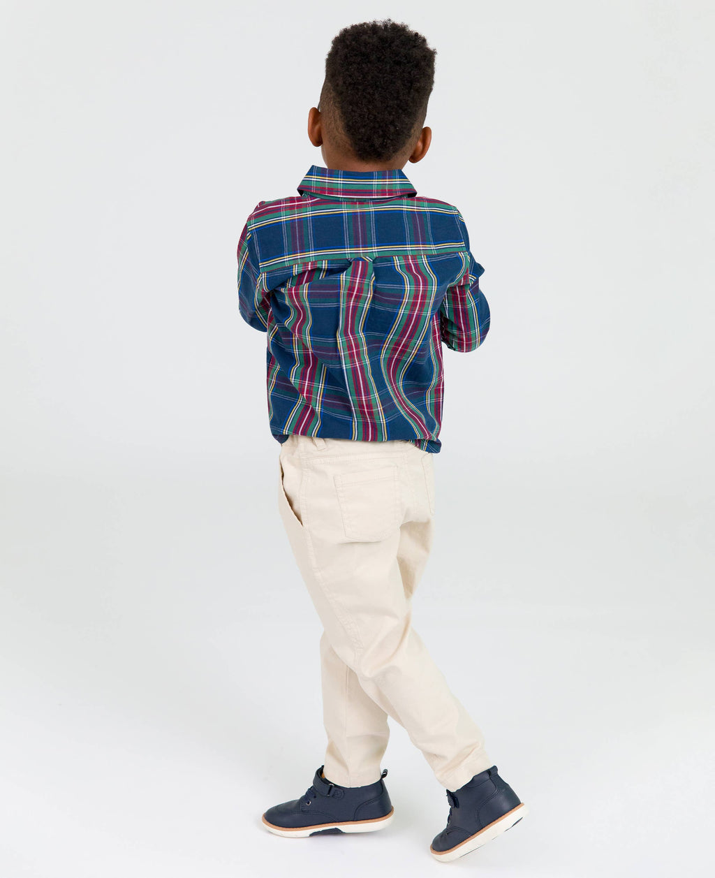 Young boy wearing RuffleButts + RuggedButts - Boys Khaki Stretch Chino Pants: Off-White paired with a blue and red plaid shirt and navy shoes, standing against a white background in a curated children’s boutique style.