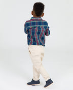 Young boy wearing RuffleButts + RuggedButts - Boys Khaki Stretch Chino Pants: Off-White paired with a blue and red plaid shirt and navy shoes, standing against a white background in a curated children’s boutique style.