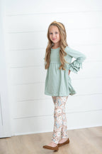 Young girl wearing a light green top and patterned pants standing against a white wooden panel background.