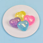 Four glittery heart-shaped squishies in yellow, pink, purple, and blue, arranged on a small white plate against a light blue background.