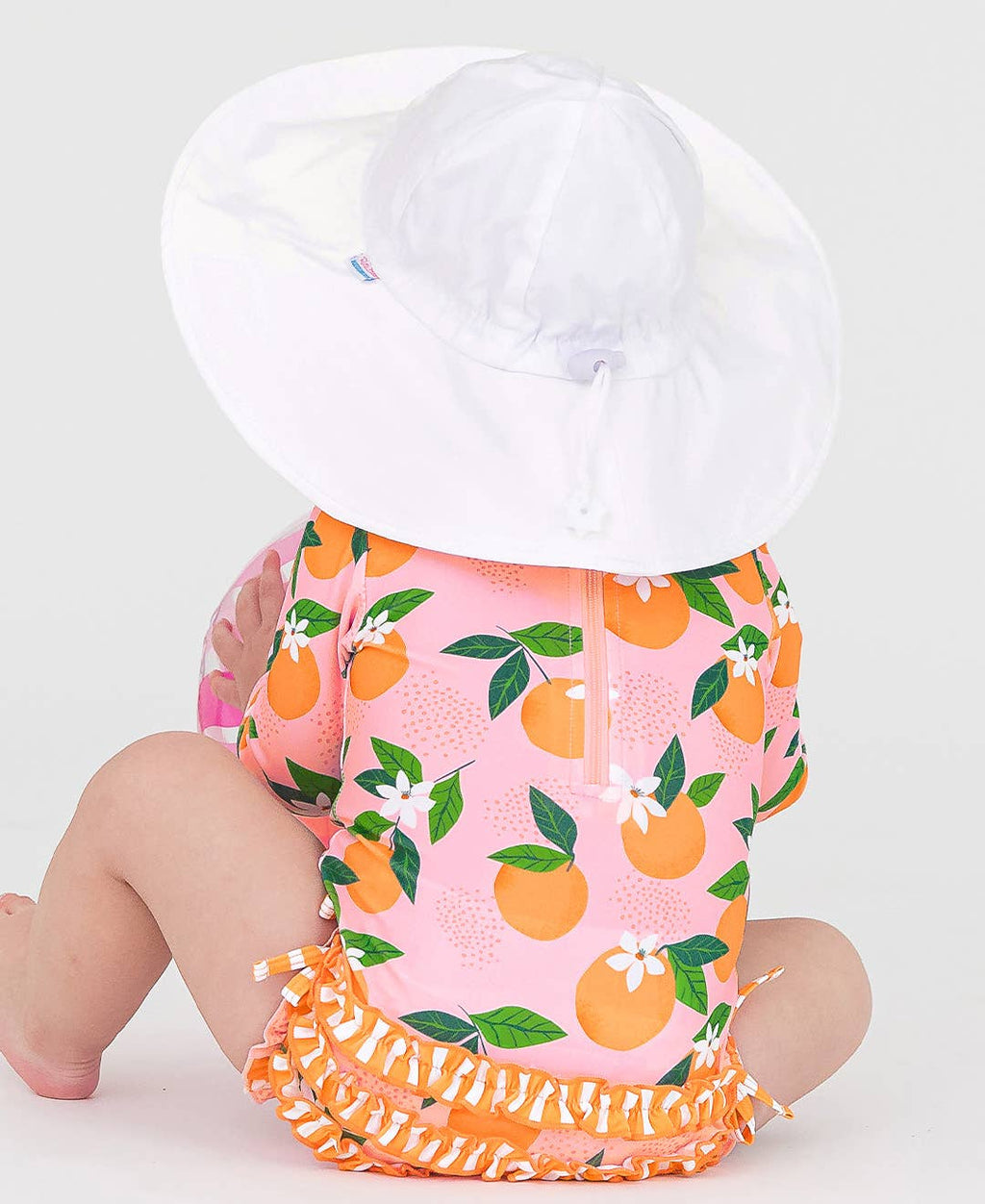 Baby wearing a bright orange and pink outfit with fruit patterns and ruffles, paired with a large white RuffleButts + RuggedButts - Kids Sun Protective Hat, showcasing stylish kids clothing.