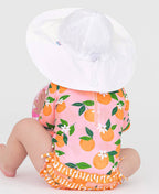 Baby wearing a bright orange and pink outfit with fruit patterns and ruffles, paired with a large white RuffleButts + RuggedButts - Kids Sun Protective Hat, showcasing stylish kids clothing.