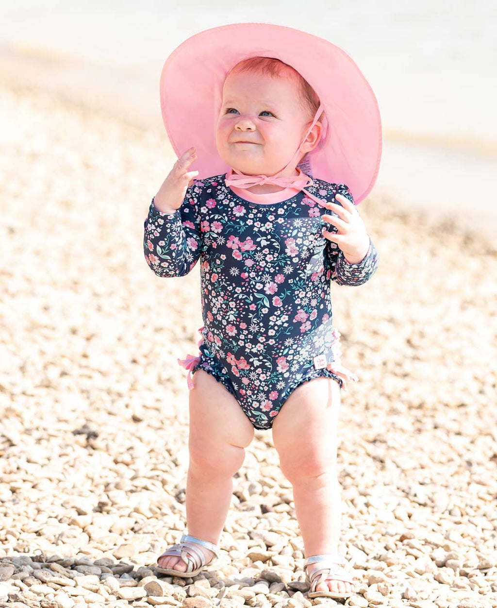 Baby standing on a pebble beach wearing a floral swimsuit and a pink RuffleButts + RuggedButts - Kids Sun Protective Hat, perfect for stylish kids clothing and sun protection.