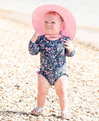 Baby standing on a pebble beach wearing a floral swimsuit and a pink RuffleButts + RuggedButts - Kids Sun Protective Hat, perfect for stylish kids clothing and sun protection.