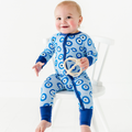 Baby wearing Kiki + Lulu - Mamma Mia! Here, I Go Again Convertible Footies with blue circular patterns, sitting on a white chair and holding a wooden teether in a bright, cozy setting.