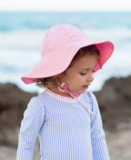 Toddler wearing a pink RuffleButts + RuggedButts - Kids Sun Protective Hat by the beach, paired with a soft blue and white striped outfit, perfect for stylish kids clothing and sun safety.