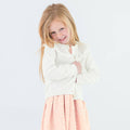 Little girl wearing the RuffleButts + RuggedButts - Girls Off-White Sweater Knit Ruffle Trim Cardigan: White, paired with a light pink polka dot skirt, smiling shyly against a white background.