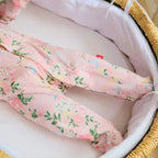 A close-up of a baby's legs wearing light pink footed pajamas with a green and yellow floral print, resting inside a white bassinet.
