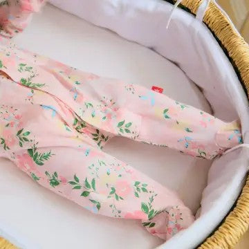 A close-up of a baby's legs wearing light pink footed pajamas with a green and yellow floral print, resting inside a white bassinet.