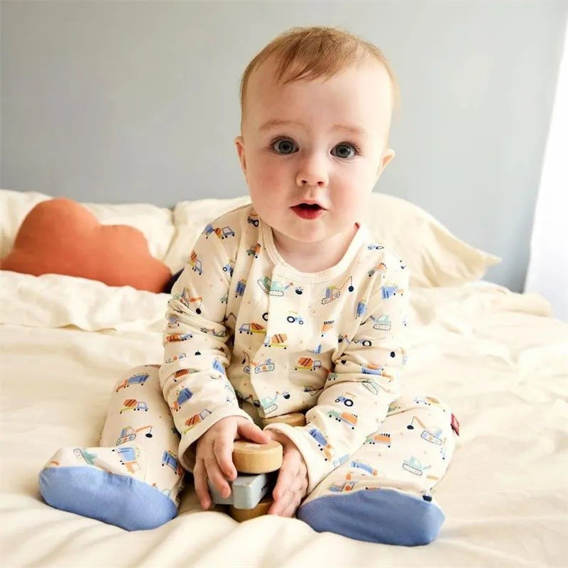 A baby sitting on a bed wearing a cream-colored long-sleeve footed pajama set with a colorful cartoon car pattern and blue socks, holding a small wooden toy.