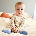 A baby sitting on a bed wearing a cream-colored long-sleeve footed pajama set with a colorful cartoon car pattern and blue socks, holding a small wooden toy.