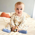 A baby sitting on a bed wearing a cream-colored long-sleeve footed pajama set with a colorful cartoon car pattern and blue socks, holding a small wooden toy.